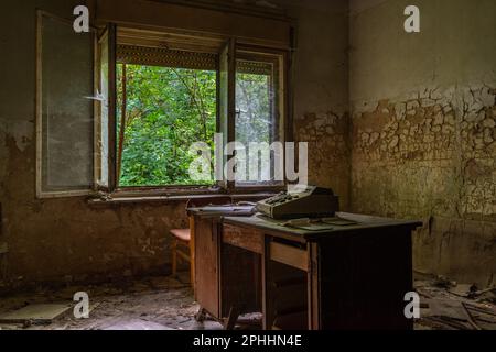 Vieille machine à écrire sur un bureau en bois dans une pièce en ruines et abandonnée, avec une vue sur la nature qui a surgrandi l'environnement urbain Banque D'Images