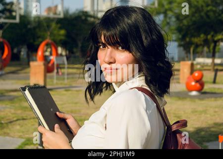 jeune femme latine, vénézuélienne, étudiant à l'université avec des vêtements décontractés et un sac à dos, tenant un livre, sérieuse regardant l'appareil photo à l'extérieur. Banque D'Images