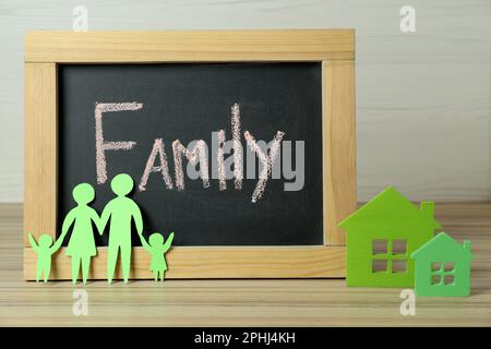 Bonne journée en famille. Tableau noir, parents en papier et leurs enfants près des modèles de maison sur une table en bois Banque D'Images