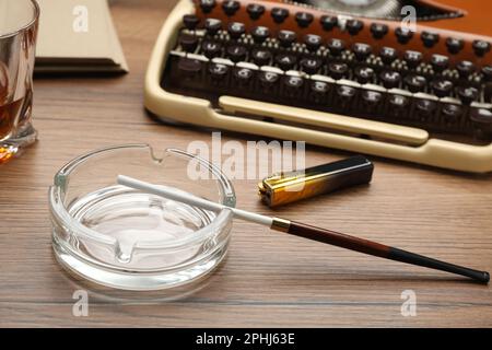 Cendrier en verre avec porte-cigarettes long et briquet près d'une machine à écrire vintage sur une table en bois Banque D'Images