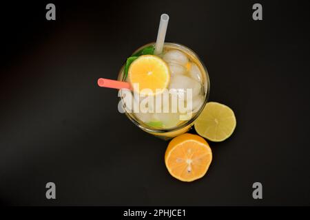 Un grand verre à facettes avec un mélange de jus d'agrumes fraîchement pressés avec de la glace sur fond noir, à côté de tranches de citron et d'orange. Vue de dessus, fl Banque D'Images