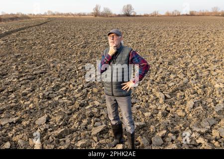 Un agriculteur âgé inquiet se tenant dans un champ labouré et regardant le ciel pour les prévisions météorologiques Banque D'Images