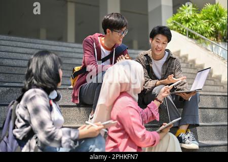Un groupe d'étudiants de divers horizons asiatiques parlent, se détendent et travaillent ensemble sur leurs travaux scolaires dans les escaliers devant le bâtiment du campus Banque D'Images