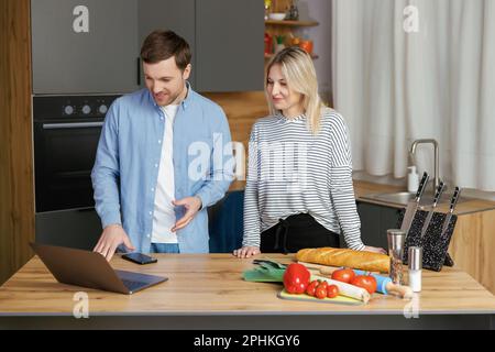 Un agréable couple familial se tenant près d'une grande table en bois dans une cuisine moderne, regardant l'écran d'un ordinateur portable. Un jeune couple heureux commence à préparer le déjeuner Banque D'Images