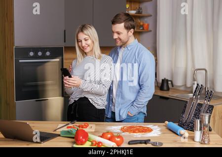 Un couple souriant et aimant prépare une salade ensemble tout en se tenant dans une cuisine à la maison et en utilisant un téléphone portable. Couple mignon homme et femme prenant la photo de foo Banque D'Images