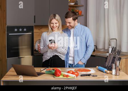 Un couple souriant et aimant prépare une salade ensemble tout en se tenant dans une cuisine à la maison et en utilisant un téléphone portable. Couple mignon homme et femme prenant la photo de foo Banque D'Images