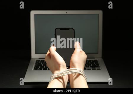 Femme avec des mains liées tenant le smartphone près de l'ordinateur portable sur fond sombre, gros plan. Dépendance à l'Internet Banque D'Images