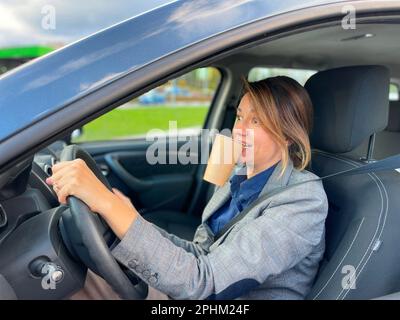 vue latérale d'une femme d'affaires conduisant une voiture en ville avec la circulation tenant une tasse de café dans la bouche avec les yeux grands regardant la route avec le visage surpris Banque D'Images