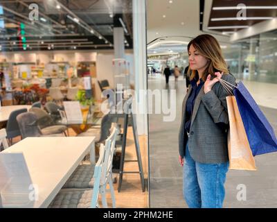 Jeune caucasienne attrayante femme debout à l'intérieur dans le centre commercial regardant la vitrine avec des meubles de maison. Jolie table et chaises de choix pour femme Banque D'Images