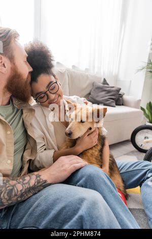 Tatoué homme embrassant positive afrique copine américaine avec le chien handicapé à la maison, image de stock Banque D'Images