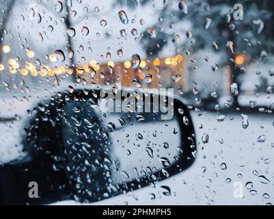 Gros plan des gouttelettes d'eau sur la vitre latérale de la voiture pendant la neige en hiver, sur fond de rétroviseur et de bokeh des feux de rue. Banque D'Images