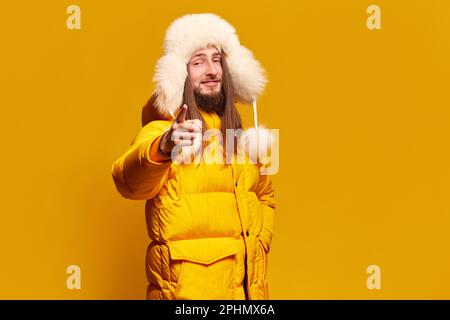 Portrait d'un jeune homme heureux et souriant en hiver manteau chaud et chapeau de fourrure posé sur fond jaune studio Banque D'Images
