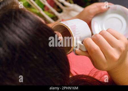 vue de dessus arrière d'une femme à peau blanche non reconnaissable assise à la maison, buvant du café dans une petite tasse. Banque D'Images