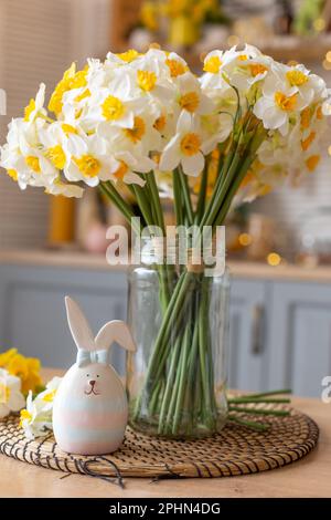 Une jolie figurine en céramique de lapin se trouve sur la table de cuisine à côté d'un vase en verre avec des fleurs de jonquilles. Banque D'Images