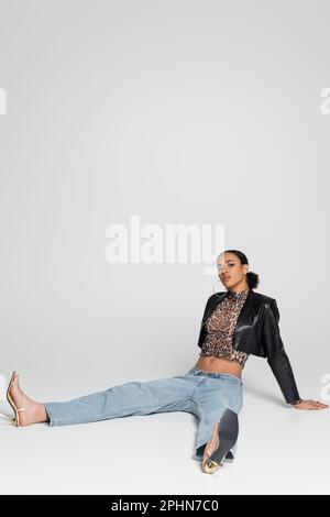 pleine longueur de jeune afro-américaine dans des sandales à talons hauts et une tenue tendance assise sur gris, image de stock Banque D'Images