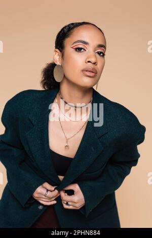 jeune modèle afro-américain avec maquillage posé dans le blazer bleu foncé de laine isolé sur beige, image de stock Banque D'Images