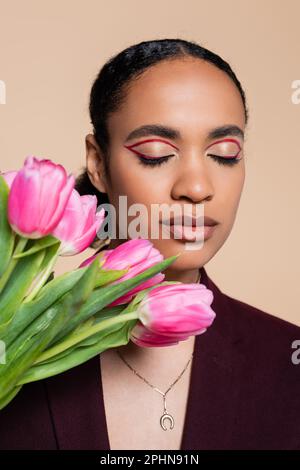 charmante femme afro-américaine avec des yeux fermés posant avec des tulipes roses isolées sur beige, image de stock Banque D'Images