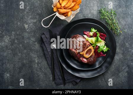 Steaks Osso Buco. Bœuf grillé Osso Buco ou steak de viande de portreuse avec garniture de salade et de frites sur un panneau de marbre noir sur du vieux bois Banque D'Images