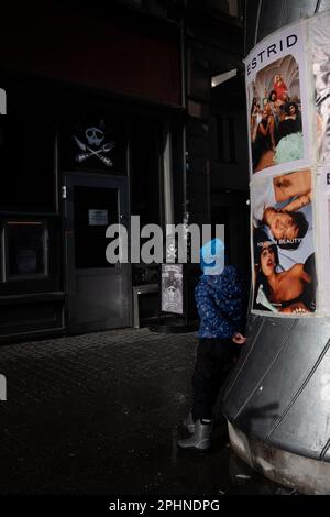 Une personne en imperméable bleu est debout sur le trottoir adjacent à un poteau de rue avec des affiches Banque D'Images