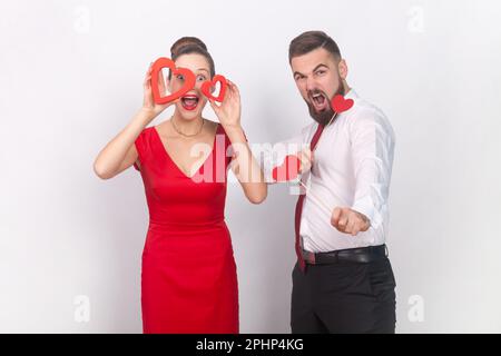 Portrait de l'homme fou en chemise blanche et femme stupéfait en robe rouge debout ensemble, tenant des figures de coeur, étant émotive. Prise de vue en studio isolée sur fond gris. Banque D'Images