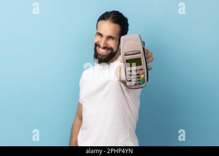 Portrait d'un homme joyeux avec une barbe portant un T-shirt blanc montrant le terminal de pos, vous suggérant d'utiliser les paiements sans contact. Studio d'intérieur isolé sur fond bleu. Banque D'Images