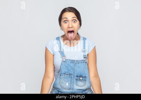 Portrait d'une femme folle désobéissante portant une combinaison en denim qui colle la langue et grimace, montrant une expression désolante et désolante. Prise de vue en studio isolée sur fond gris. Banque D'Images