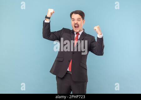 Portrait d'un homme très heureux et excité debout avec des poings serrés, célébrant sa victoire, hurlant, portant une combinaison noire avec une cravate rouge. Prise de vue en studio isolée sur fond bleu clair. Banque D'Images