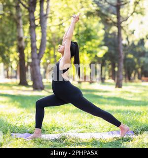 Jeune femme enceinte menant un mode de vie sain et pratiquant le yoga, exécute l'exercice Virabhadrasana 1, posture du guerrier, s'entraîne dans le parc sur une wa Banque D'Images