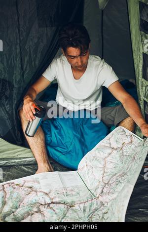 Jeune homme regardant une carte des sentiers de montagne planifiant le prochain voyage en vacances d'été assis dans une tente sur le camping. Concept de vie de camp Banque D'Images