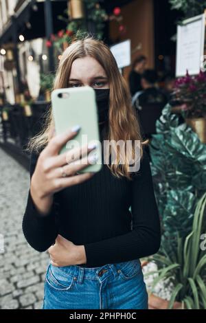 Jeune femme ayant un appel vidéo parlant en marchant en centre-ville portant le masque facial pour éviter l'infection virale et pour empêcher la propagation de la maladie dans le TI Banque D'Images