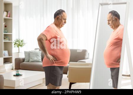 Homme mature avec un grand ventre debout devant un miroir à la maison dans un salon Banque D'Images