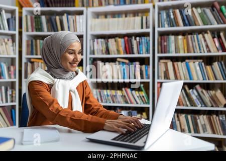 Jeune beau étudiant en hijab étudiant en ligne à distance dans la bibliothèque de l'université, femme tapant sur le clavier d'ordinateur portable à la recherche d'informations et se préparant à l'examen. Banque D'Images