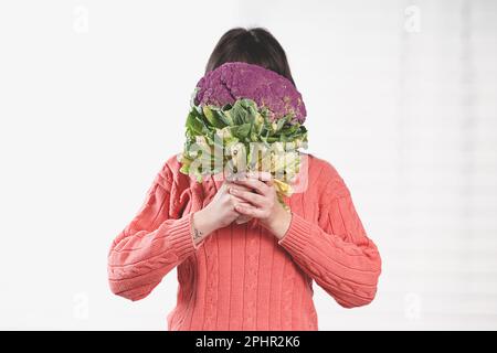Jeune caucasienne joyeuse cachant son visage derrière un chou-fleur, le tenant des deux mains, debout par une fenêtre avec un rideau blanc. Banque D'Images