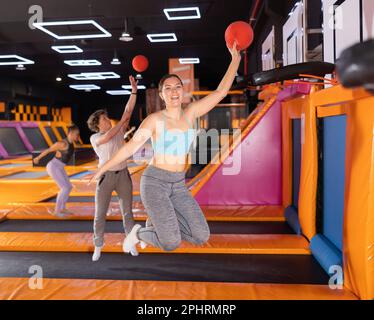 Une jeune femme européenne gaie dans des vêtements de sport jouant au basket-ball tout en rebondissant sur des trampolines colorés à l'intérieur dans le centre sportif de loisirs Banque D'Images