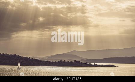 Une image de paysage monochrome dorée avec un bateau à voile sur un lac, avec les montagnes en arrière-plan et les rayons du soleil se brisant à travers un ciel nuageux s. Banque D'Images