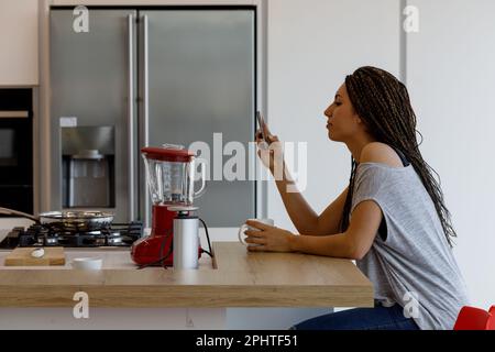 la jeune femme utilise son smartphone dans la cuisine tout en prenant le petit déjeuner ou une pause à une table d'îlot moderne et spacieuse. Appareils en acier inoxydable surrou Banque D'Images