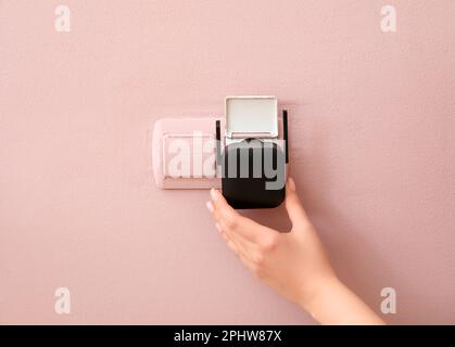 Une femme branche un répéteur WiFi noir dans la prise électrique sur un mur rose Banque D'Images