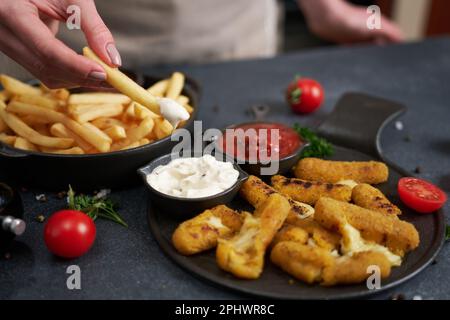 Une femme plonge les frites dans une sauce trempée avec des bâtonnets de mozzarella frits au fromage sur une table Banque D'Images