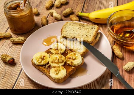 Beurre d'arachide ou pâte d'arachide dans un pot ouvert et étaler sur un sandwich toast avec banane et miel et arachides dans des coquilles dispersées sur la table Banque D'Images