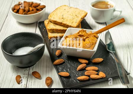 Dans un bol sur la table, le beurre d'amande biologique cru avec une cuillère en bois et des amandes éparpillées sur la table, griller le pain et le café Banque D'Images