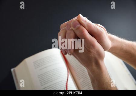 Homme religieux priant avec les mains et les doigts entremêtés et livre religieux en arrière-plan sur table noire. Vue de dessus. Banque D'Images