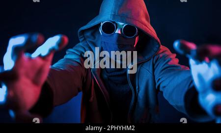 Homme effrayant en lunettes, masque et capuche. Éclairage au néon. Le jeune homme européen est dans le studio sombre Banque D'Images