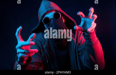 Homme effrayant en lunettes, masque et capuche. Éclairage au néon. Le jeune homme européen est dans le studio sombre Banque D'Images