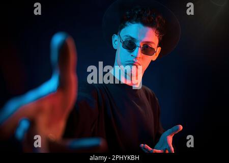 En chapeau et lunettes. Éclairage au néon. Le jeune homme européen est dans le studio sombre Banque D'Images