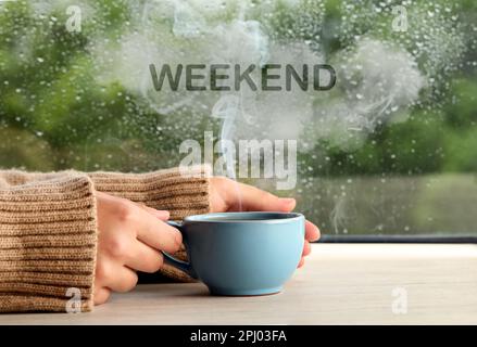 Bonne fin de semaine. Femme avec une tasse de boisson chaude à une table en bois près de la fenêtre le jour de la pluie, gros plan Banque D'Images