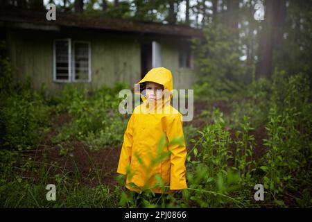 Un enfant dans un manteau jaune debout dans les bois en face d'une ancienne maison en bois. Un garçon de six ans, Pologne, Europe Banque D'Images