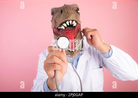 Médecin avec masque T Rex tenant un stéthoscope en gros plan sur un fond rose isolé. mise au point sélective Banque D'Images