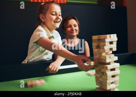 Fille excitée jouant jenga jeu avec sa mère dans la salle de jeu. Fille en retirant un bloc de la pile et en la plaçant sur le dessus de la tour. Blocs de bois empilés. G Banque D'Images