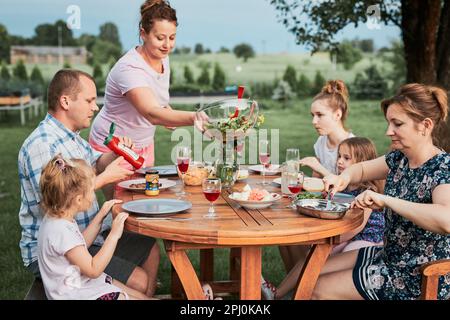 Famille ayant un repas du grill pendant le pique-nique d'été dîner en plein air dans un jardin de maison. Gros plan des personnes assises à une table avec de la nourriture et de la vaisselle Banque D'Images