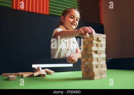 Petite fille jouant à jenga dans la salle de jeux. Tour de bâtiment pour enfants avec blocs de bois. Empilage de pièces en bois. Jeu de compétence et de plaisir Banque D'Images